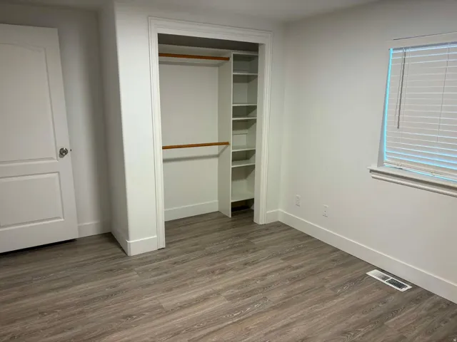 Room featuring wood-finish flooring, a built-in closet with shelving and hanging rods, a paneled door, and a window with blinds