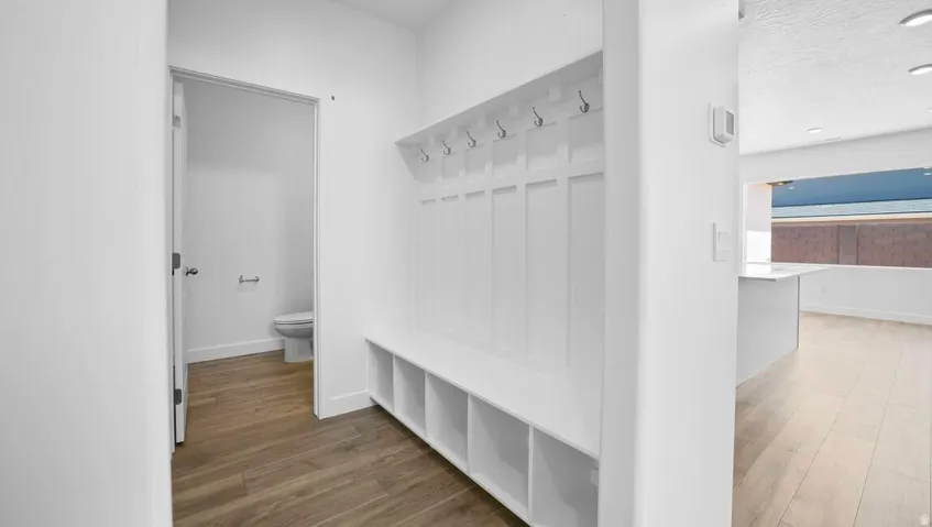 Built-in mudroom bench with storage cubbies and coat hooks, wood-finish flooring, white walls, and a powder room with a toilet and toilet paper holder