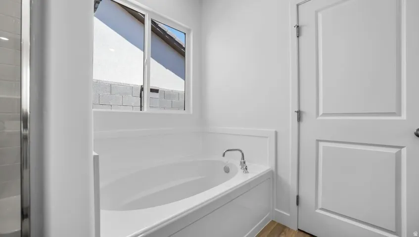 Soaking tub with a deck-mounted faucet, white wall paneling, a white paneled door, and wood-finish flooring