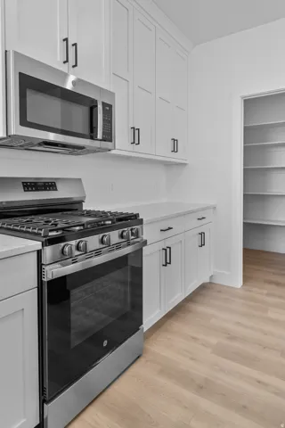 White shaker-style cabinetry with matte black hardware, stainless steel appliances, light-toned wood-finish flooring, and a walk-in pantry with built-in shelving