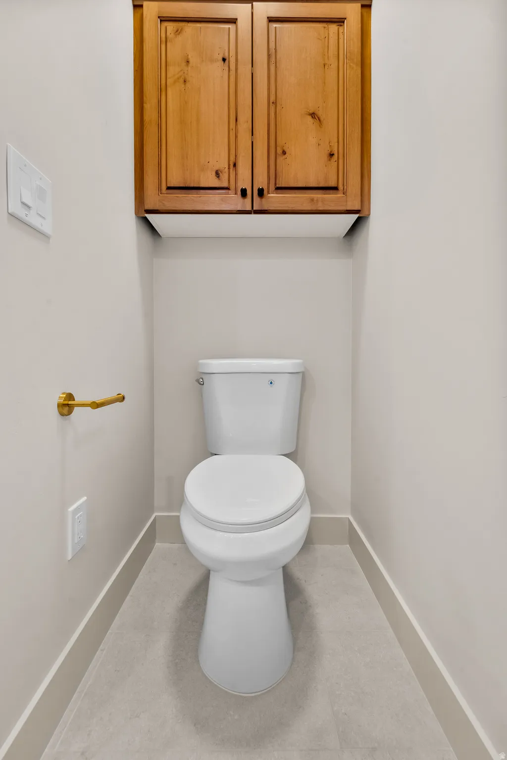 Recessed toilet with overhead wood cabinetry, gold-finish hardware, and tiled flooring