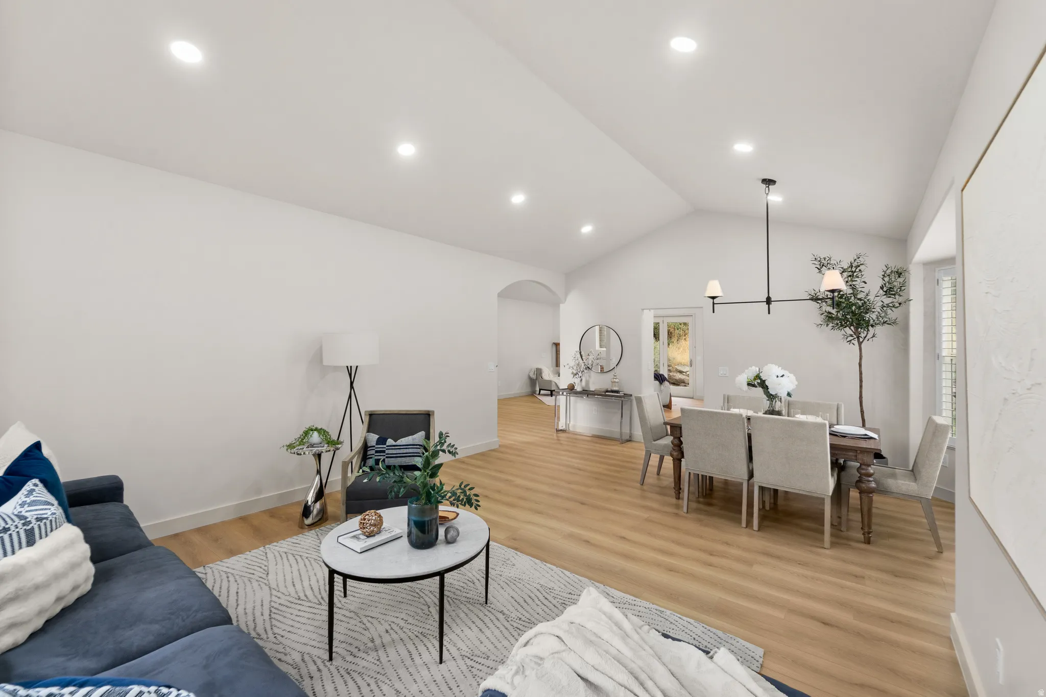 Vaulted ceilings, recessed lighting, and wood-finish flooring characterize this open-concept space, featuring an arched doorway and a dining area with a chandelier