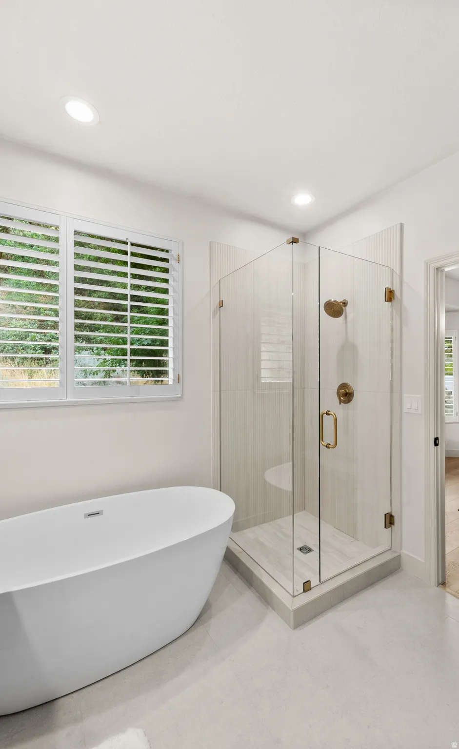 Freestanding bathtub, glass-enclosed shower with gold-tone fixtures, white plantation shutters, recessed lighting, and light-toned floor tiling