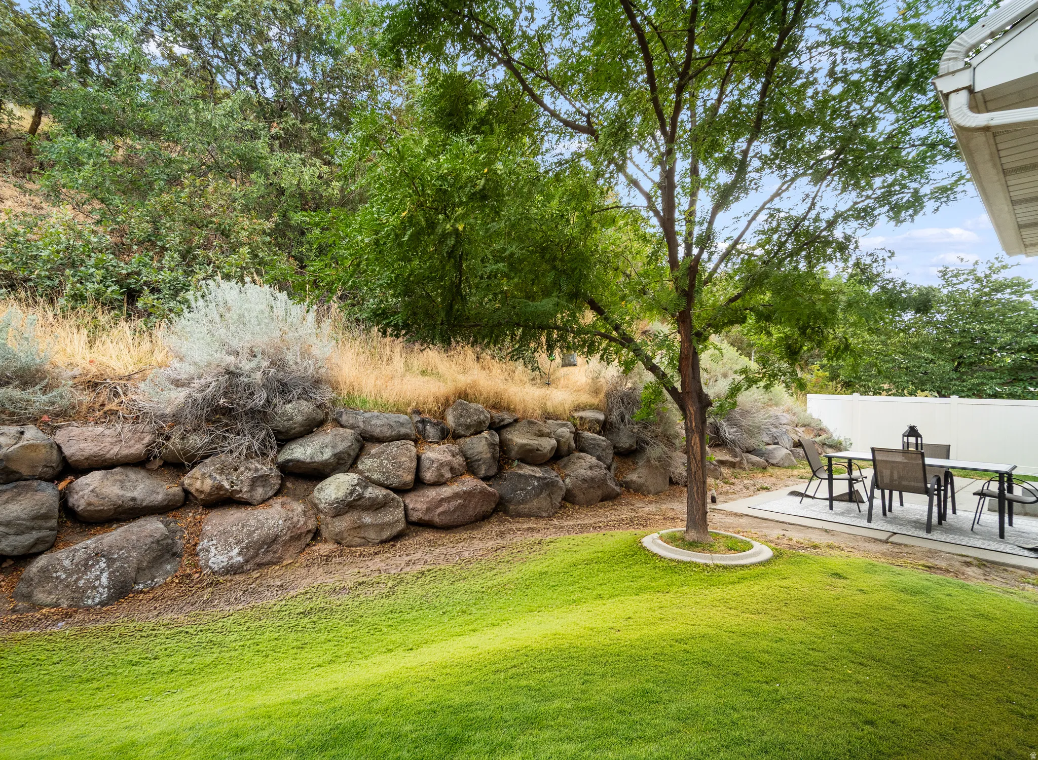 Expansive lawn with mature landscaping, a natural rock retaining wall, and a paved patio area