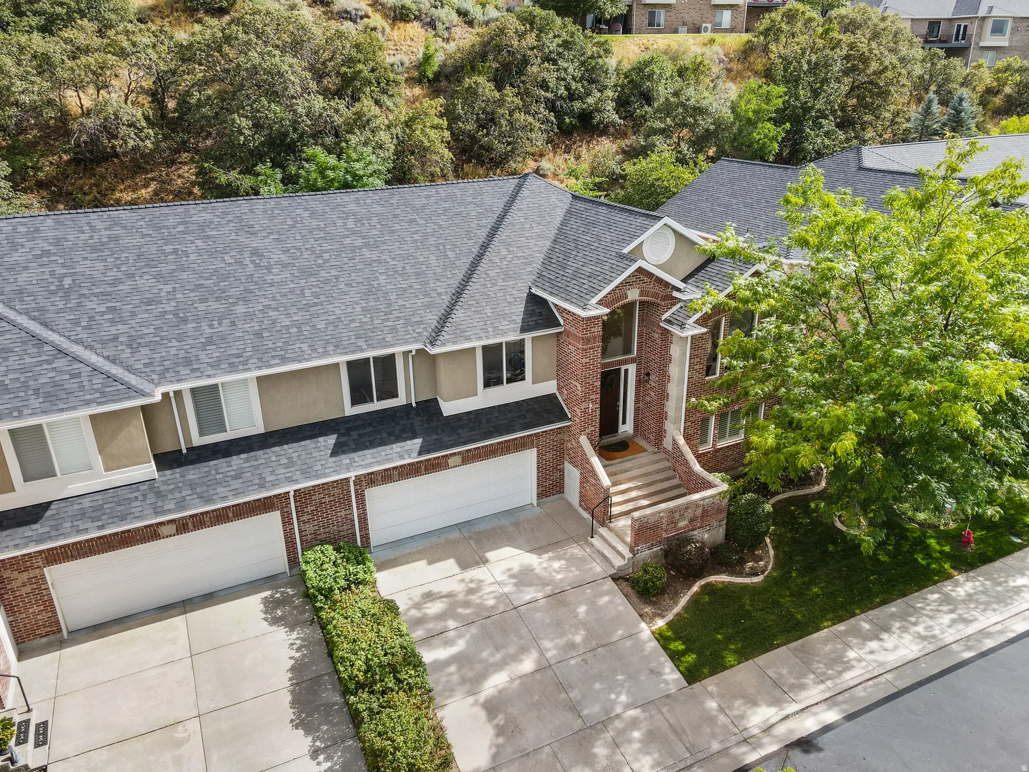 Brick and stucco exterior, multi-car garage, mature landscaping, concrete driveway, and asphalt shingle roof