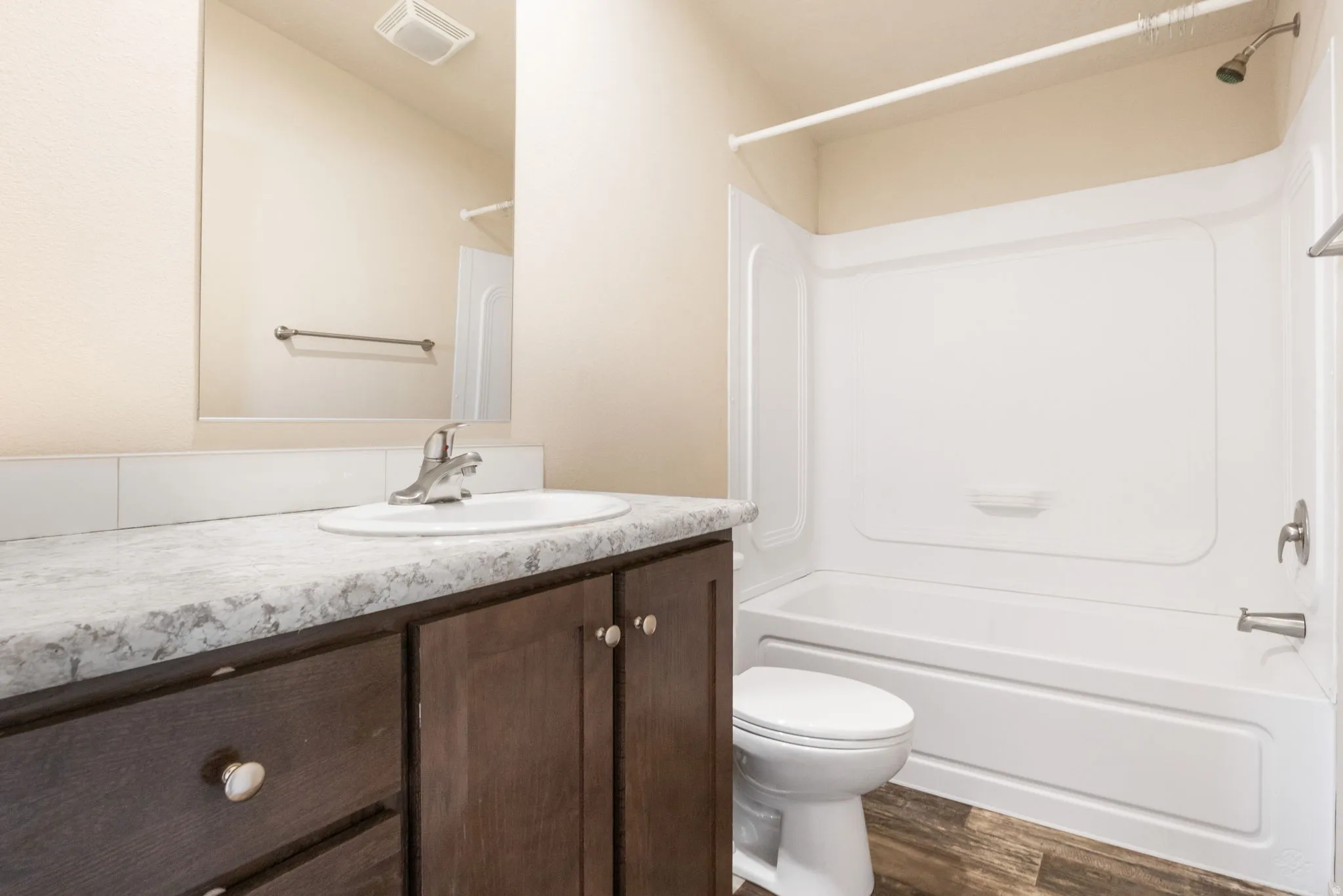 Wood-finish flooring, dark wood-tone vanity, gray-toned countertop, white drop-in sink, and a white bathtub with shower