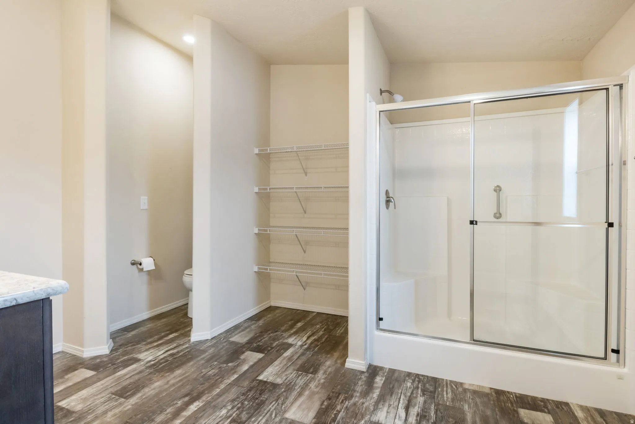 Walk-in shower with sliding glass doors, built-in shelving, wood-finish flooring, and a private commode area