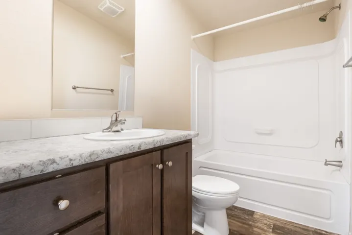 Wood-finish flooring, dark wood-tone vanity, gray-toned countertop, white drop-in sink, and a white bathtub with shower
