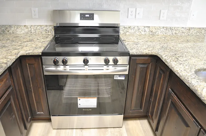 Stainless steel range, light-toned granite countertops, crisp white tile backsplash, dark wood-finish cabinetry, and wood-finish flooring