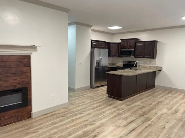 Open-concept living area with wood-finish flooring, a fireplace featuring a wood surround, a kitchen island with a stone countertop, dark wood cabinetry, and stainless steel appliances