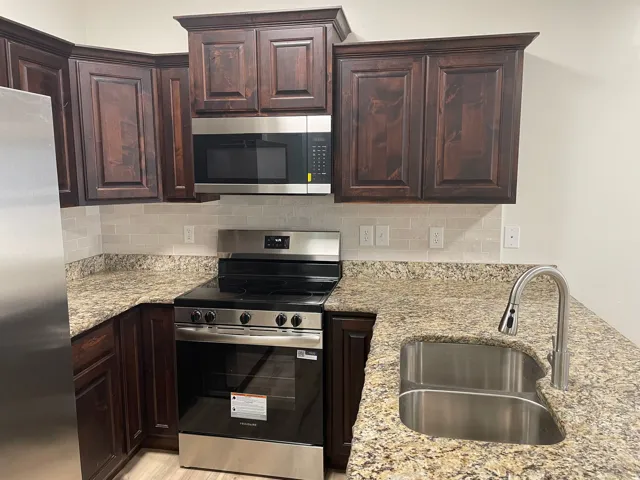 Rich wood cabinetry, stainless steel appliances, speckled stone countertops, subway tile backsplash, and a stainless steel double-basin sink with a gooseneck faucet