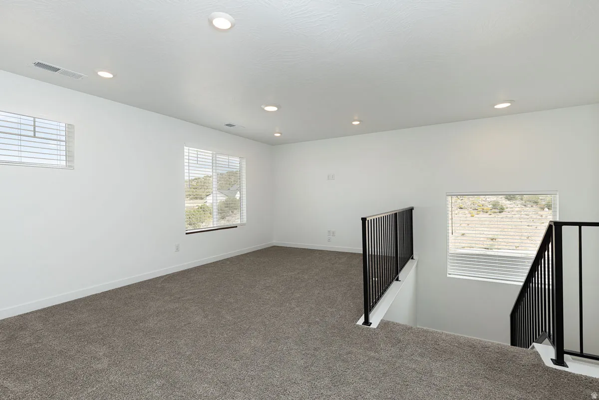 Recessed ceiling lighting, multiple windows with blinds, dark carpet flooring, and black metal stair railings
