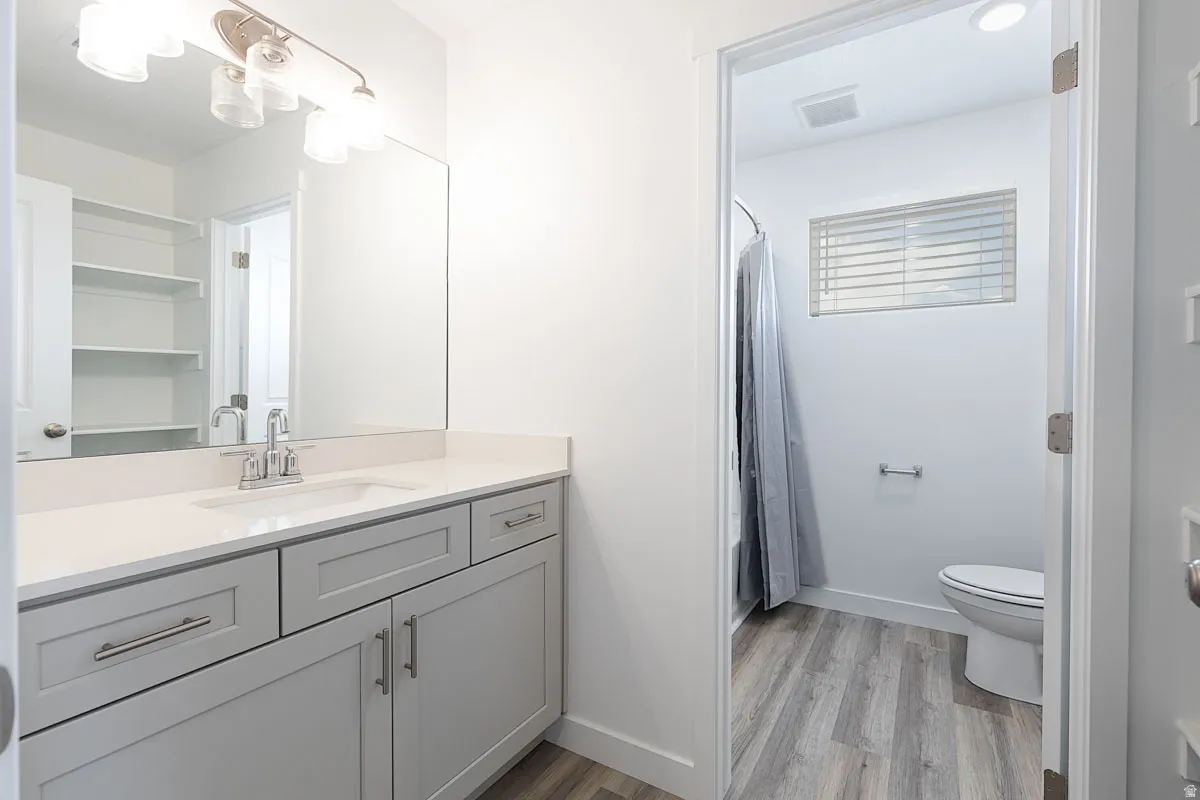 Bathroom with wood-finish flooring, a vanity featuring shaker-style cabinetry and a light-toned countertop, an integrated sink with a chrome faucet, a large mirror, and built-in shelving
