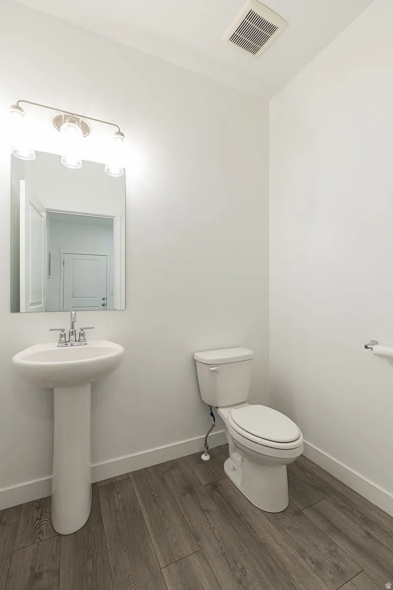 Pedestal sink, wall-mounted mirror, overhead vanity lighting, toilet, and wood-finish flooring