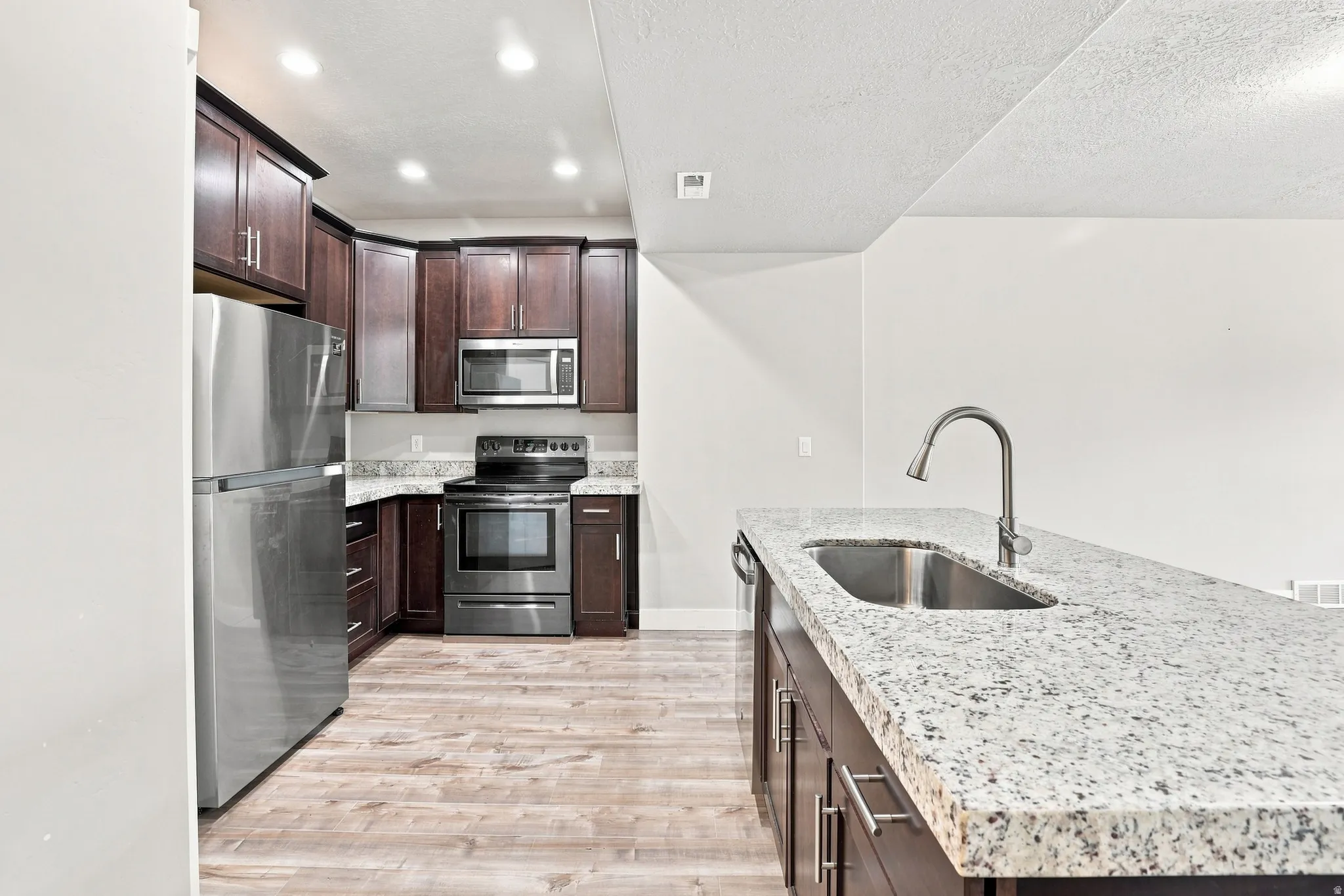 Kitchen featuring wood-finish flooring, dark wood cabinetry, stainless steel appliances, and granite countertops, with an integrated kitchen island sink