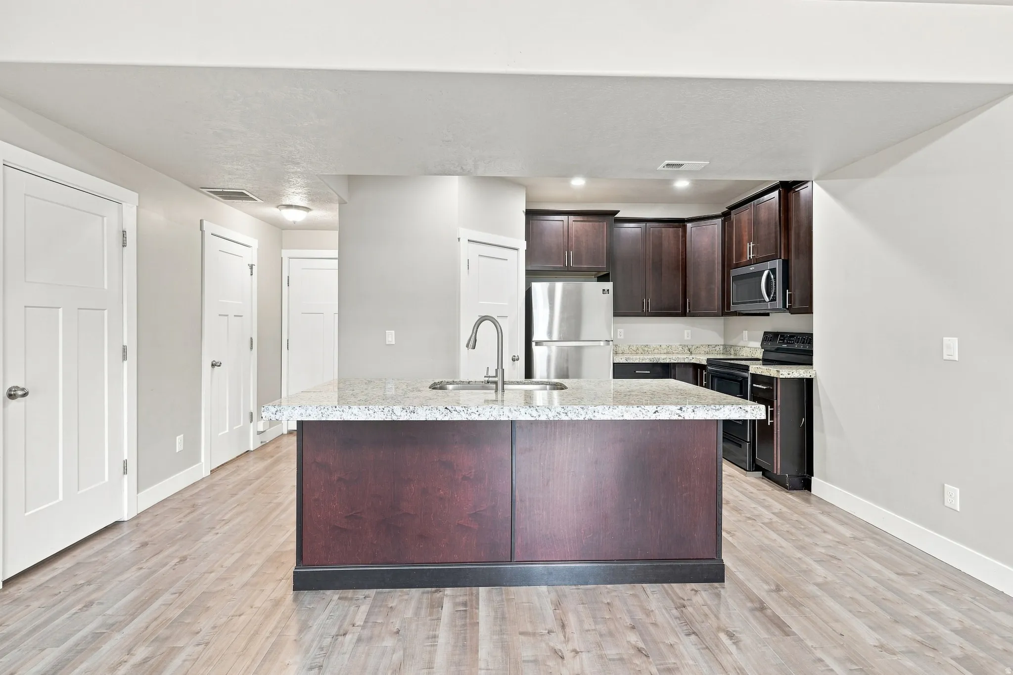 Kitchen island with a light-colored countertop and integrated sink, stainless steel appliances, dark wood-finish cabinetry, and wood-finish flooring