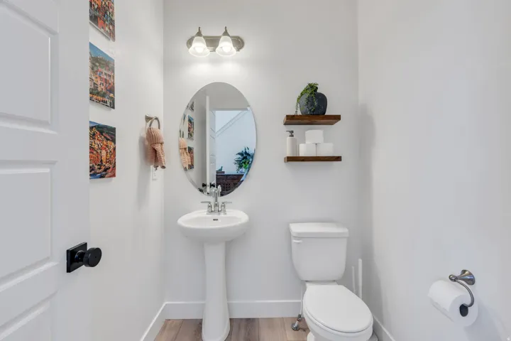 Pedestal sink with an oval mirror, wall-mounted light fixture, and wood-finish floating shelves