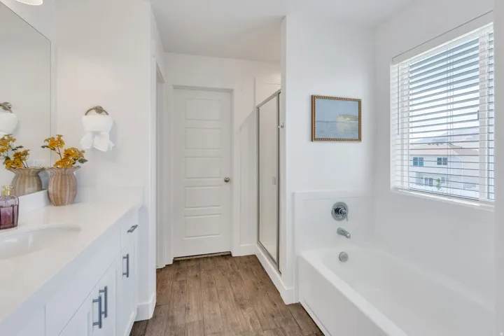 Wood-finish flooring, white vanity with integrated sink, white bathtub, enclosed shower, and a window with blinds