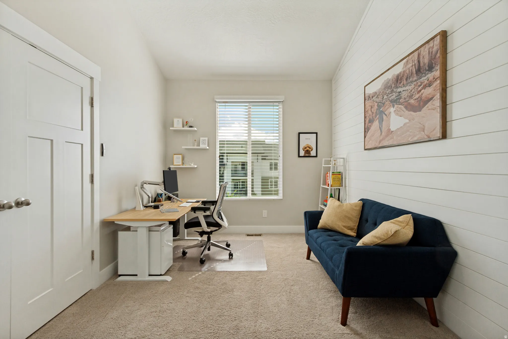 Room featuring a window with blinds, carpeted flooring, a white paneled accent wall, and a solid white door with a three-panel design