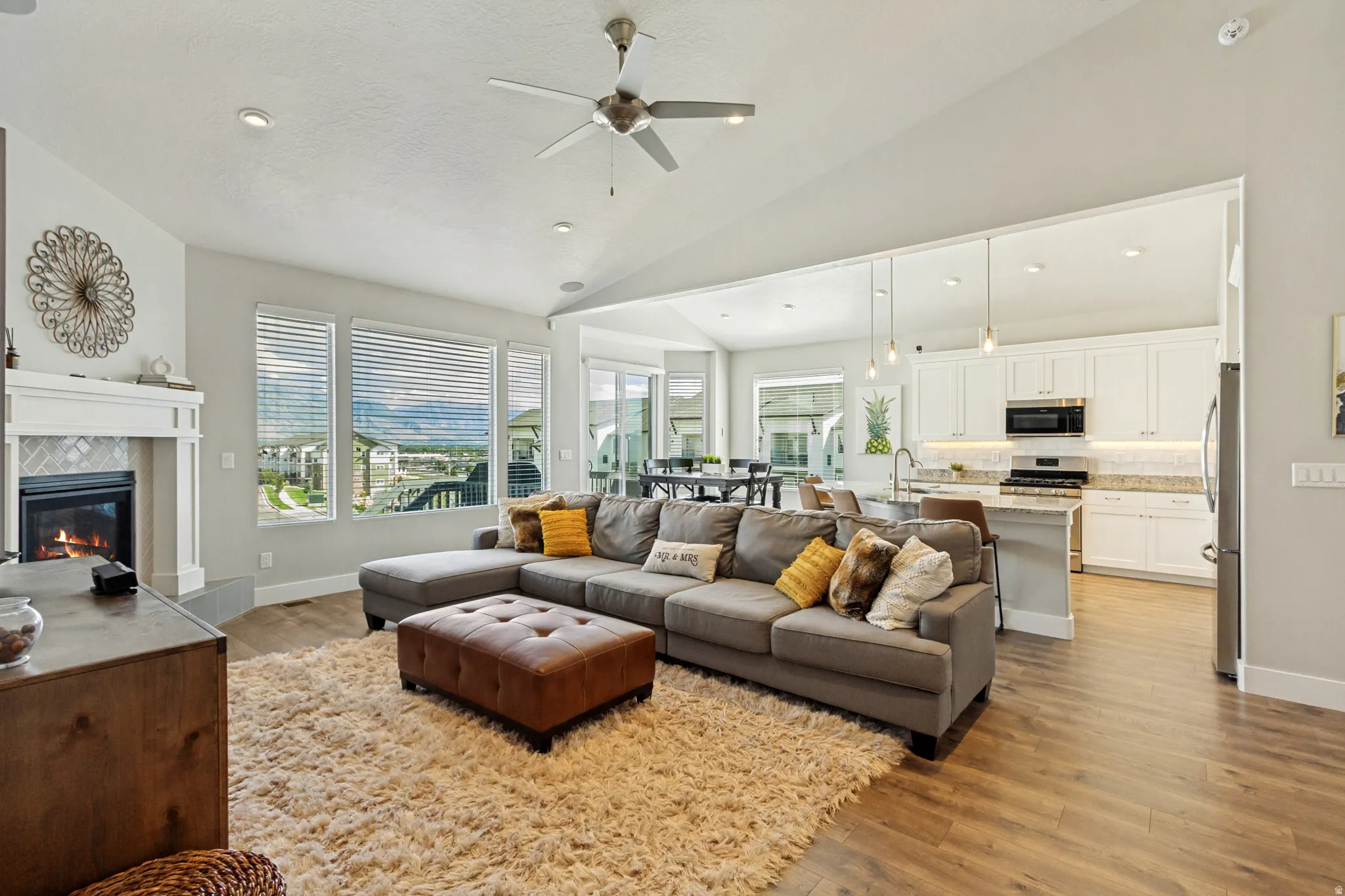 Vaulted ceiling with a ceiling fan, fireplace with a tile surround and mantel, wood-finish flooring, and a kitchen with white cabinetry and stainless steel appliances