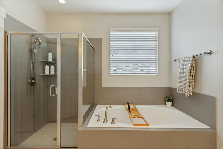 Recessed bathtub with deck-mounted fixtures, separate glass-enclosed shower with tile surround, and a window with blinds