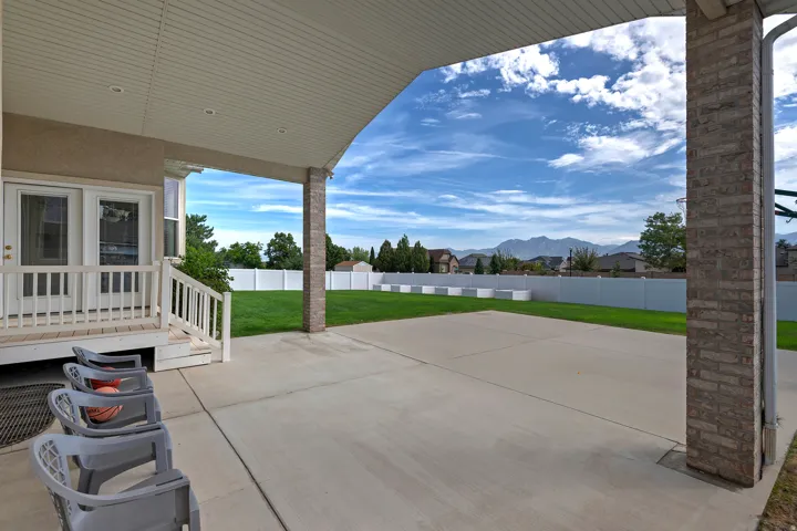 Covered patio with recessed lighting, concrete slab, brick-clad pillar, and a large grassy yard featuring a white privacy fence and distant mountains