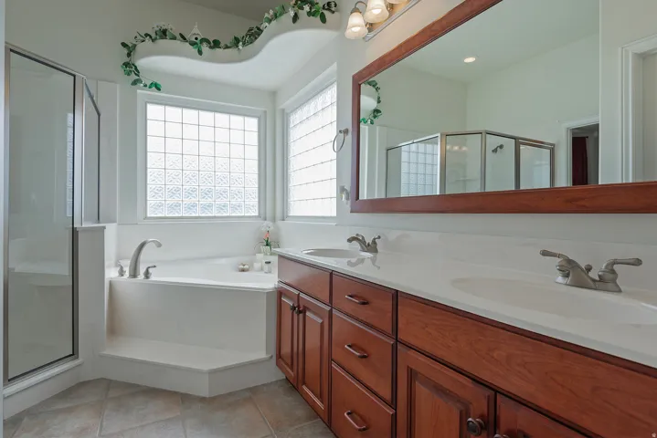 Built-in soaking tub with a glass block window, clear glass shower enclosure, dual basin vanity with wood-finish cabinetry, and a large wall-mounted mirror