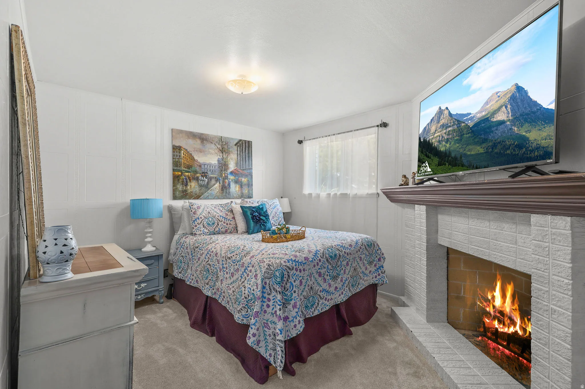 Carpeted bedroom featuring a white brick fireplace with a wood mantel, a window with sheer curtains, and a ceiling-mounted light fixture