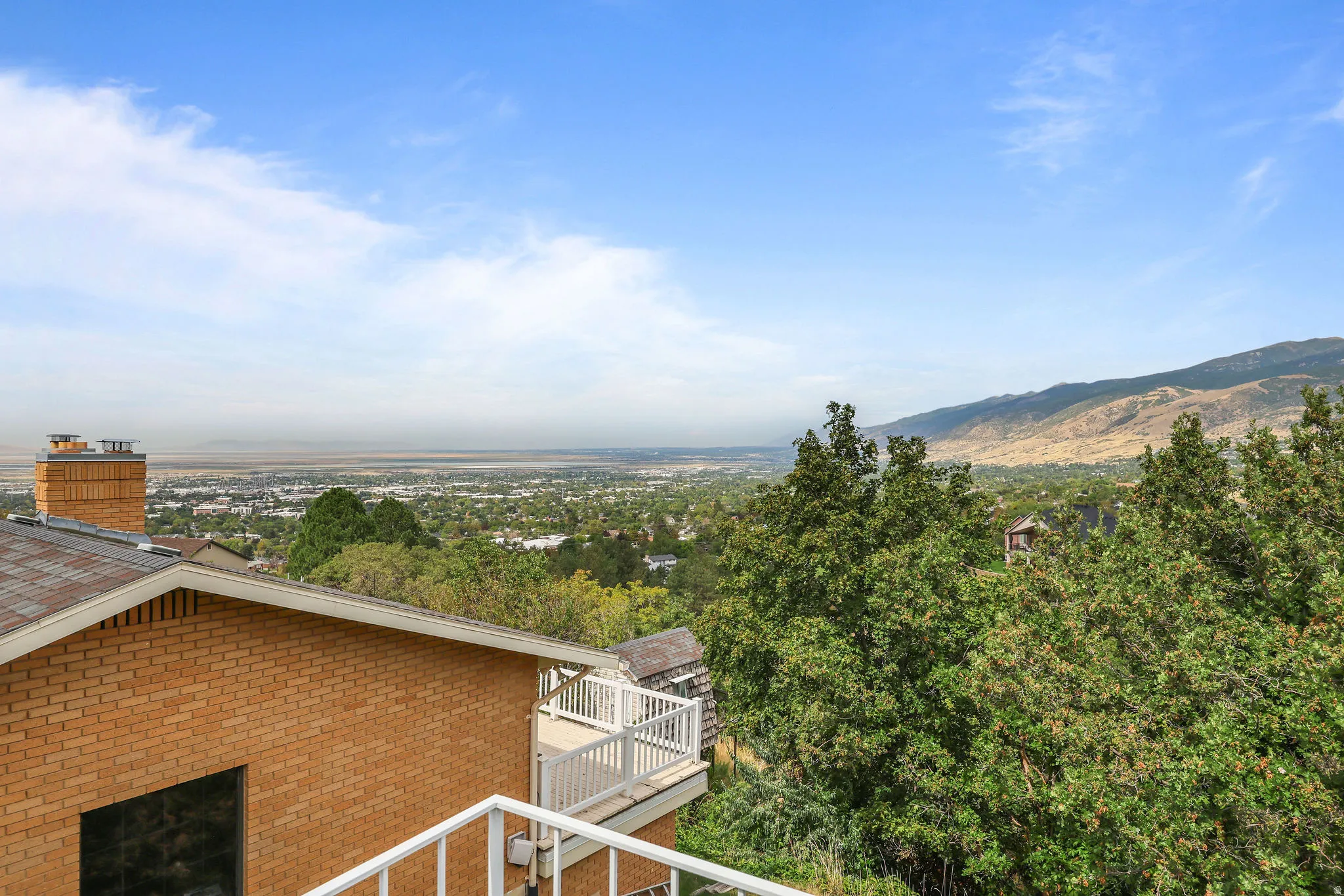 Expansive city and mountain backdrop, mature trees, brick exterior, white balcony railing, and a shingle roof