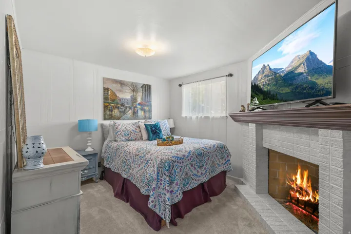 Carpeted bedroom featuring a white brick fireplace with a wood mantel, a window with sheer curtains, and a ceiling-mounted light fixture