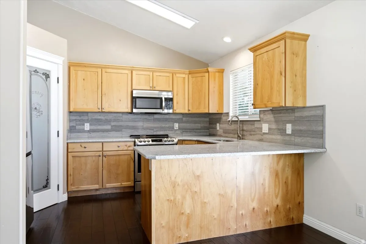 Vaulted ceiling kitchen featuring light wood cabinetry, stone-look tile backsplash, stainless steel appliances, and dark wood-finish flooring