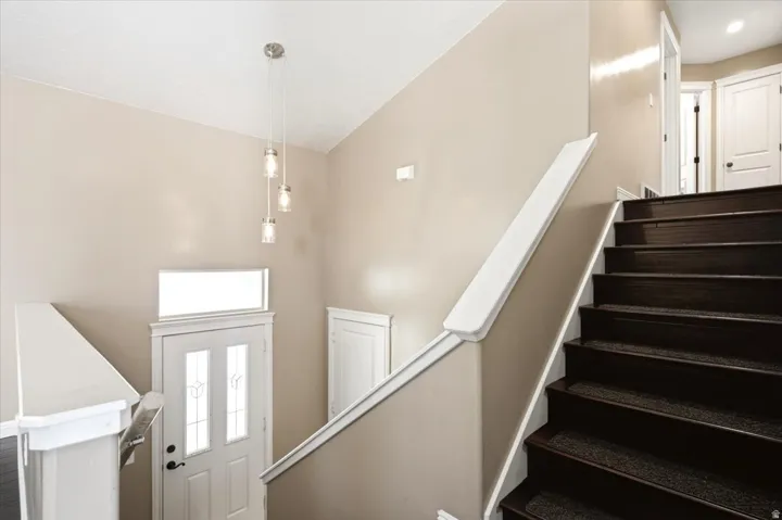 Dark wood stairs with carpeted treads, white handrail, pendant light fixture, and white entry door with decorative glass