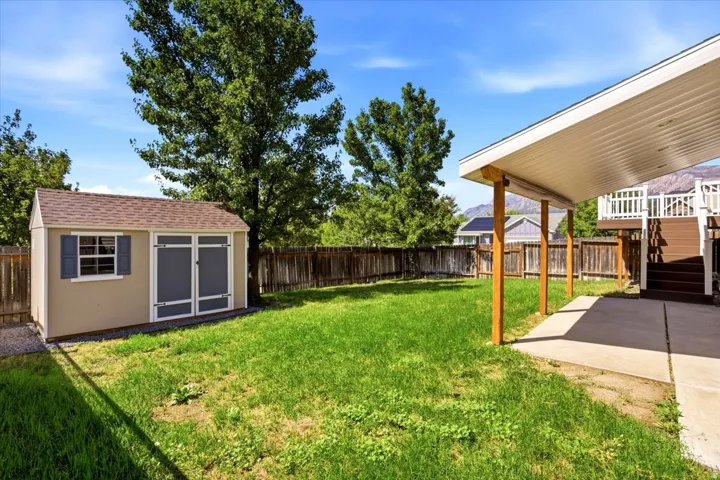 Expansive grassy backyard featuring a paved patio with a covered section supported by wood posts, a wood privacy fence, and an outdoor storage shed with shutters