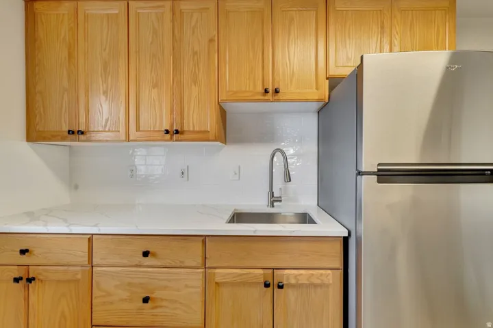 Wood-finish cabinetry with black hardware, white subway tile backsplash, white countertop with gray veining, stainless steel sink with pull-down faucet, and stainless steel refrigerator