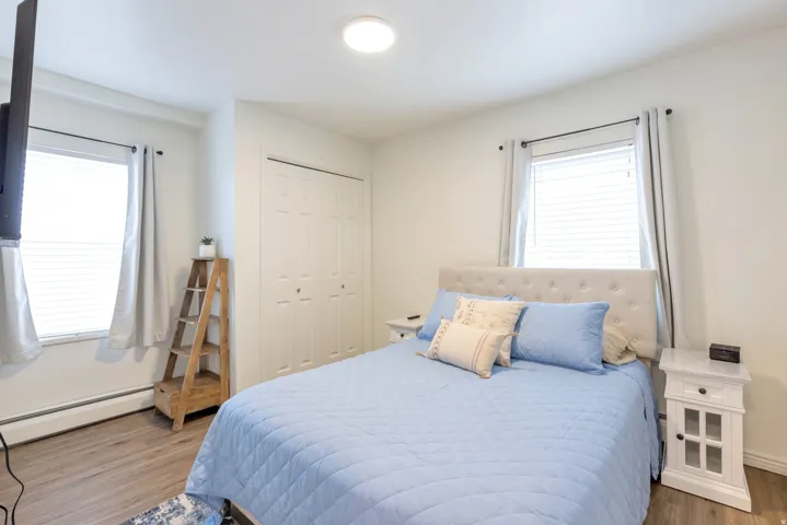 Recessed ceiling light, two windows with blinds, wood-finish flooring, bi-fold closet doors, and a padded headboard