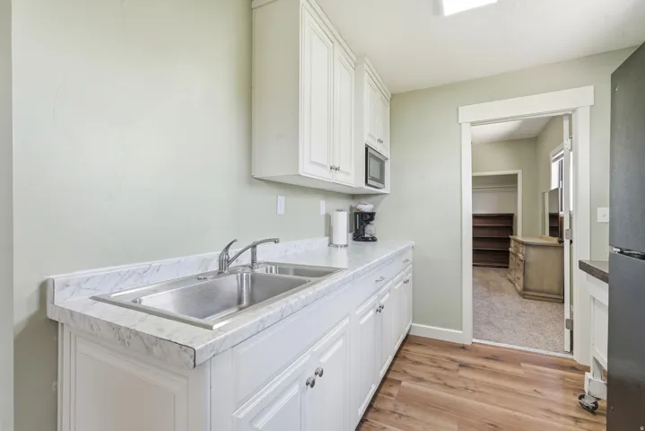 White cabinetry with silver hardware, stainless steel double basin sink, light-toned countertop, wood-finish flooring, and a built-in microwave