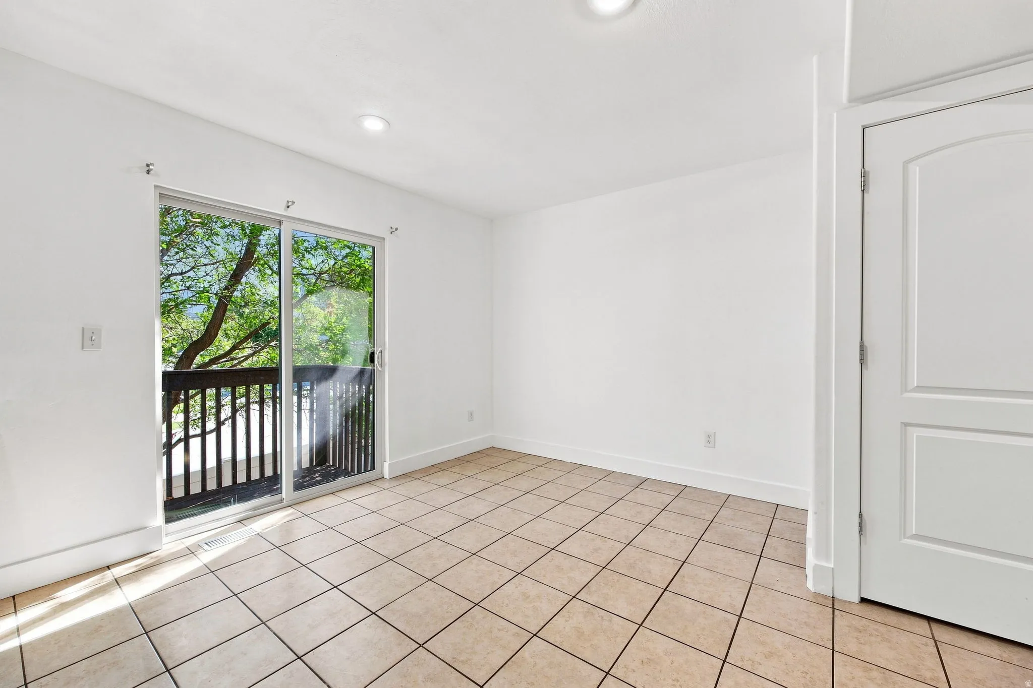 Expansive tile flooring, sliding glass door leading to a balcony with dark wood railing, and recessed lighting