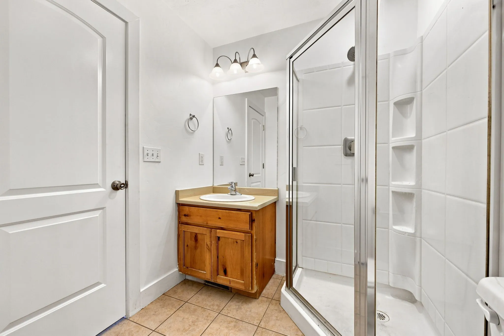 Glass-enclosed shower with integrated shelving, wood-finish vanity with countertop, and ceramic tile flooring