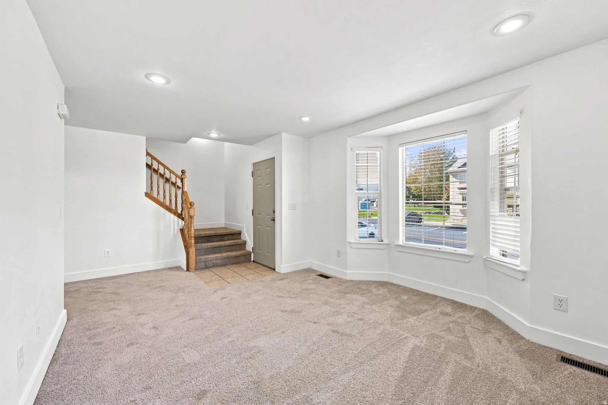 Expansive room with neutral carpeting, recessed lighting, a wood staircase, and a large bay window
