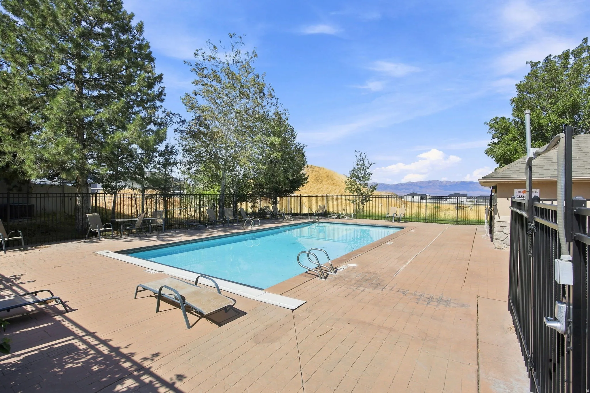Outdoor swimming pool with a surrounding paved deck, enclosed by a black metal fence, with a nearby structure featuring stone accents