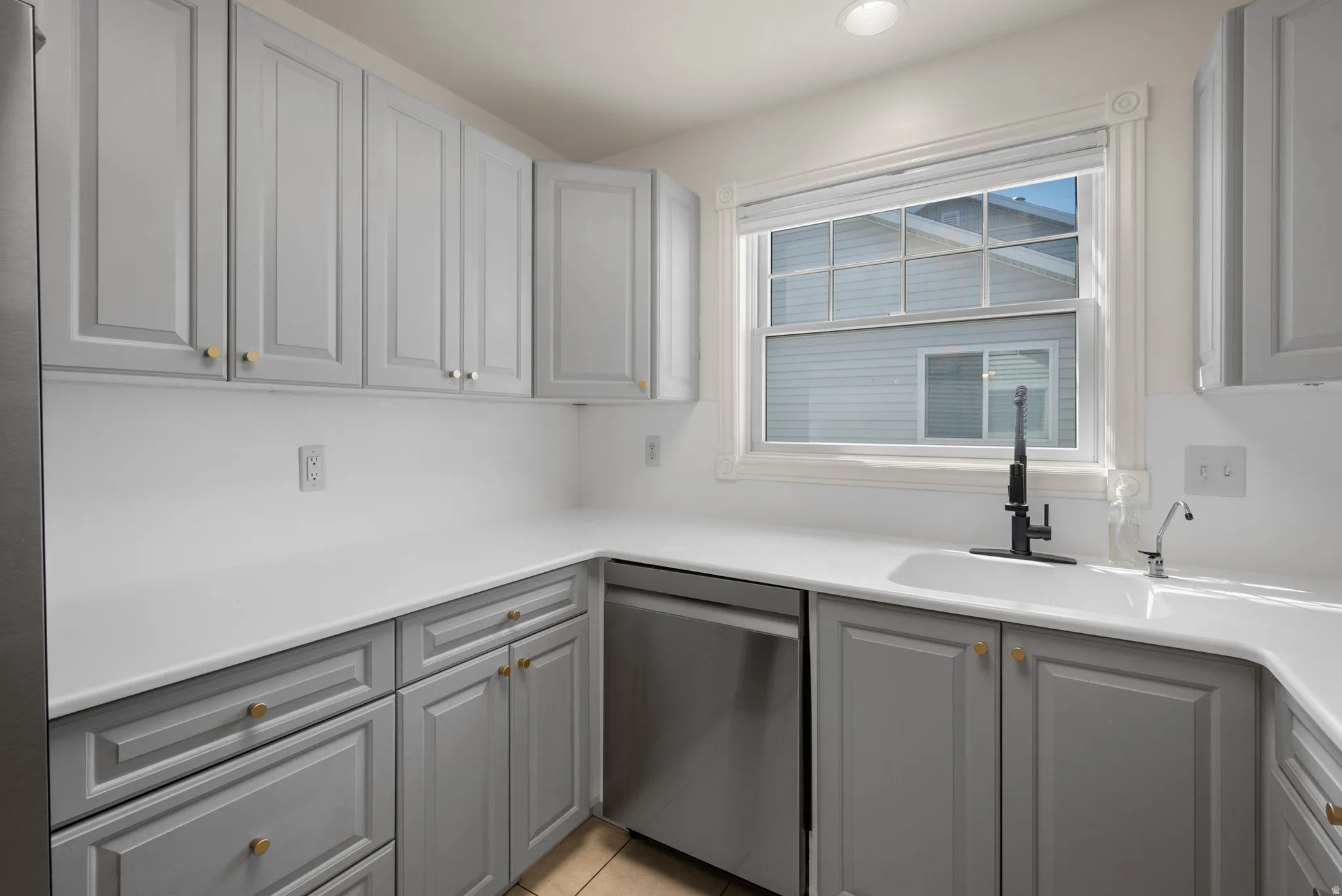 Gray shaker-style cabinetry with gold-tone hardware, white countertops, an undermount sink with a matte black faucet, and a stainless steel dishwasher