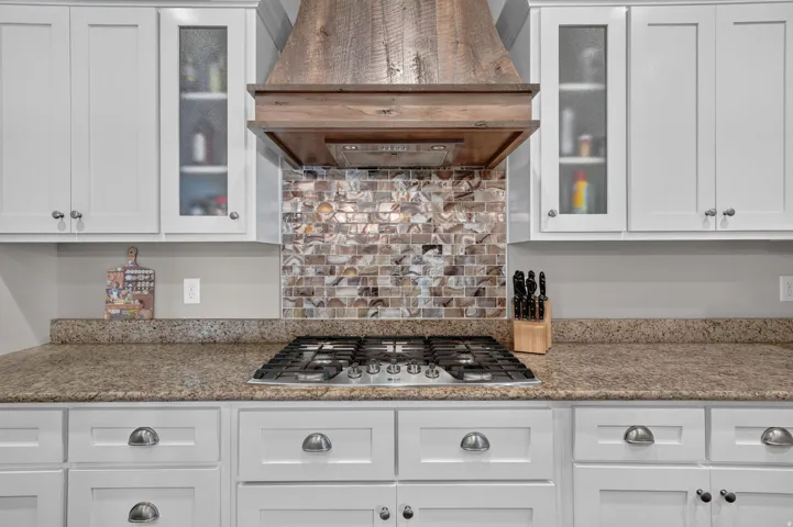 White shaker-style cabinetry with glass-front accents, mosaic tile backsplash, built-in gas cooktop, and wood-finish range hood