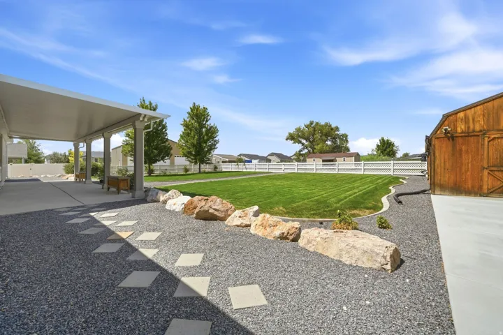 Expansive green lawn, white lattice fence, covered patio with support columns, gravel landscaping with stepping stones, and a wooden barn-style structure