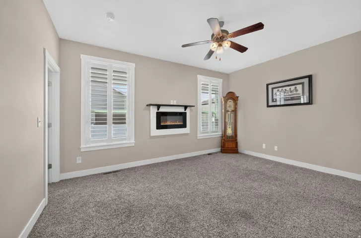 Built-in electric fireplace with mantel, ceiling fan with integrated lighting, two windows with plantation shutters, and neutral-toned wall paint
