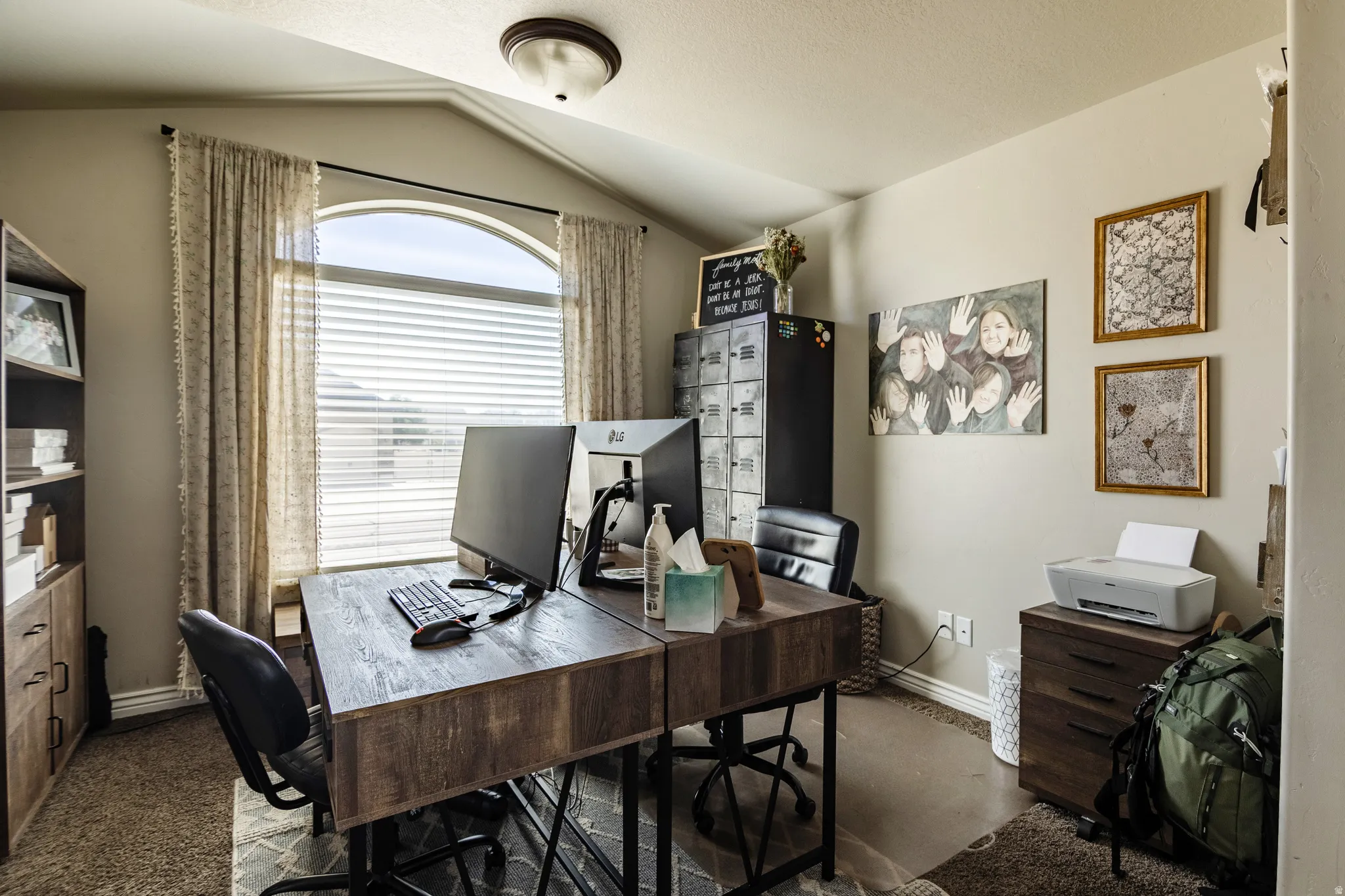 Arched window with blinds and drapes, wood-finish desk, wood-finish shelving unit, carpeted flooring, and a flush mount light fixture