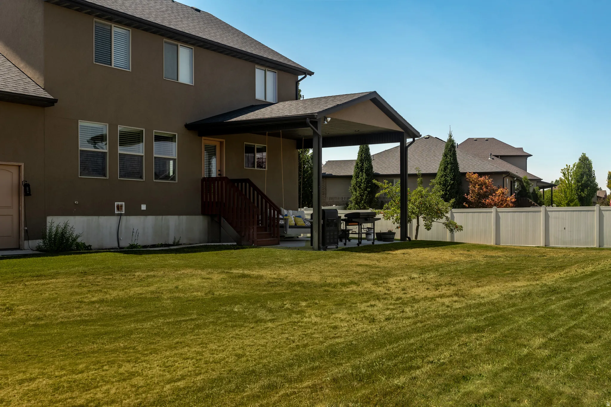 Expansive grass area, stucco exterior, covered patio, wood-finish deck with stairs, and white privacy fencing