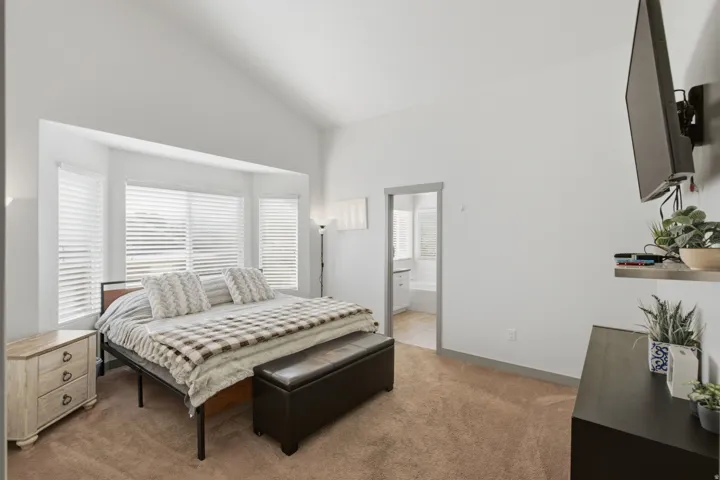 Vaulted ceiling, bay window with blinds, carpeted flooring, and an open doorway leading to an additional space