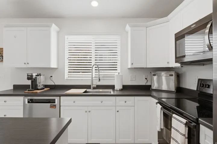 White shaker cabinetry, dark countertops, undermount stainless steel sink with a gooseneck faucet, stainless steel dishwasher, and a built-in microwave