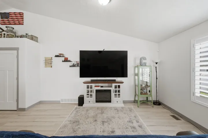 Vaulted ceiling, wood-finish flooring, white plantation shutters, white interior door with gray trim, and a built-in electric fireplace