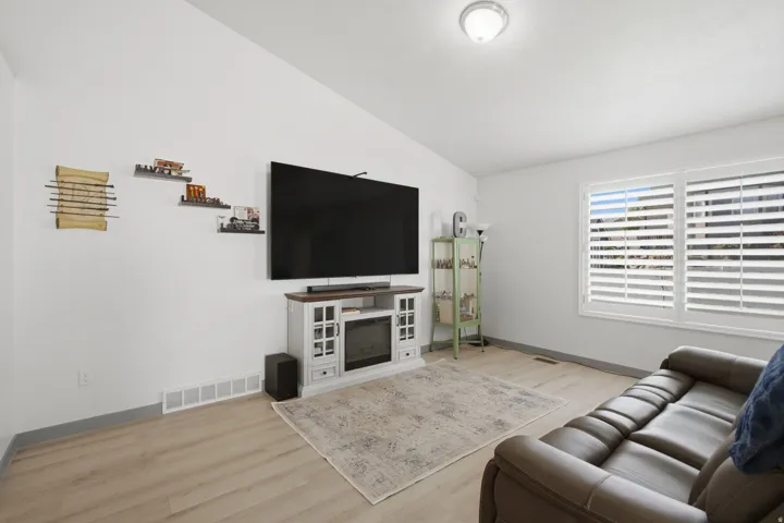 Vaulted ceiling, wood-finish flooring, white walls, built-in media console with electric fireplace, window with white plantation shutters